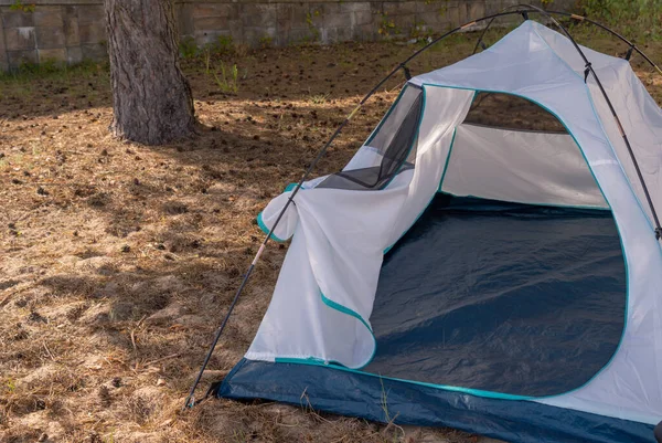 a white tourist tent is spread out on the sand under a cedar tree in the shade of a tree, the tent is open, has a green bottom, overnight stay in nature in the summer