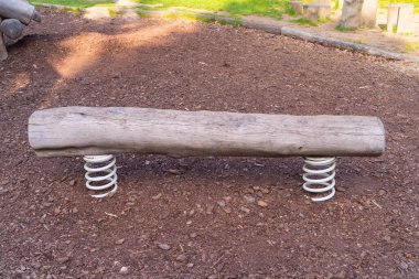 a wooden bench made of a tree trunk on metal white springs on the ground covered with shredded bark from trees, front photo, the bench is not covered with anything, dry in the park