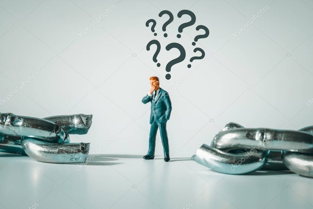Macro photo of thoughtful businessman figurine standing between broken chain. Question marks above the pondering miniature man.  Conflict, bankruptcy, indecision unemployment concept.
