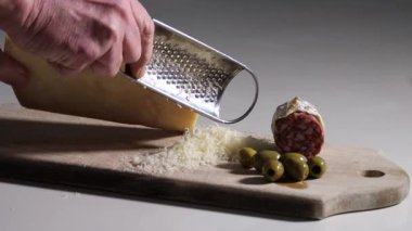 Bir insan yeşil zeytin ve salamlı ahşap kesim tahtasının üzerinde parmesan peyniri rendeliyor.. 