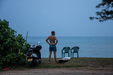 Mirissa, Sri Lanka plajda bir adam sabahın erken saatlerinde sörf tahtasıyla yol kenarında duruyor..