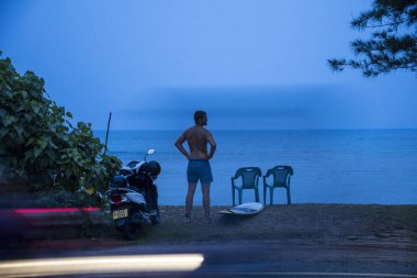 Mirissa, Sri Lanka plajda bir adam sabahın erken saatlerinde sörf tahtasıyla yol kenarında duruyor..