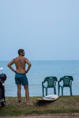 Mirissa, Sri Lanka plajda bir adam sabahın erken saatlerinde sörf tahtasıyla yol kenarında duruyor..