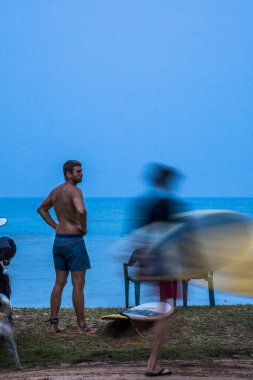 Mirissa, Sri Lanka plajda bir adam sabahın erken saatlerinde sörf tahtasıyla yol kenarında duruyor..