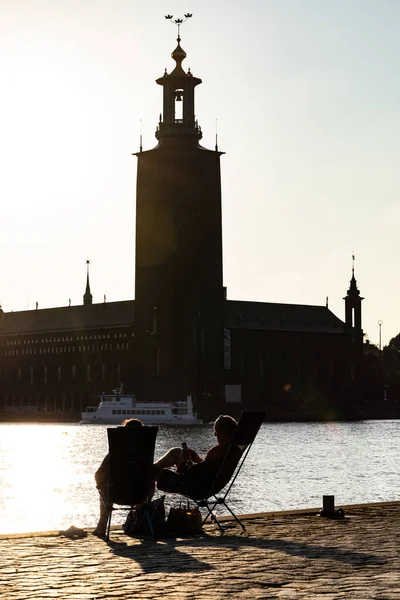 ストックホルム スウェーデンリダルフィヨルダンを見下ろす夕日のビーチチェアでリラックスしたカップルと市庁舎 — ストック写真