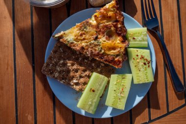 A bacon and spinach quiche is served on a plastic plastic plate with a sliced cucumber.