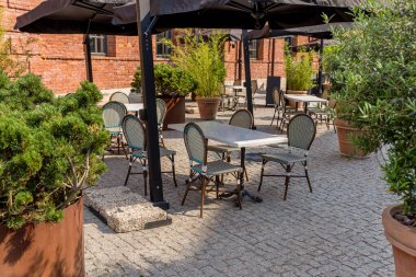 summer outdoor cafe with stone gray pavement green bushes in tubs sunny day black high umbrellas from the sun seats wrapped with artificial light rotan stone brick walls