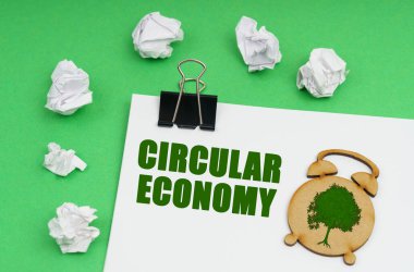 Çevresel konsept. Yeşil arka planda beyaz bir çarşaf var, üzerinde bir alarm saati figürü ve bir yazıt var - yuvarlak ekonomi, yanında buruşuk kağıt var.