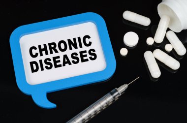 Tıbbi konsept. Siyah bir yüzeyde, bir şırınga, haplar ve üzerinde CHRONIC DISEASES yazan mavi bir çerçeve.