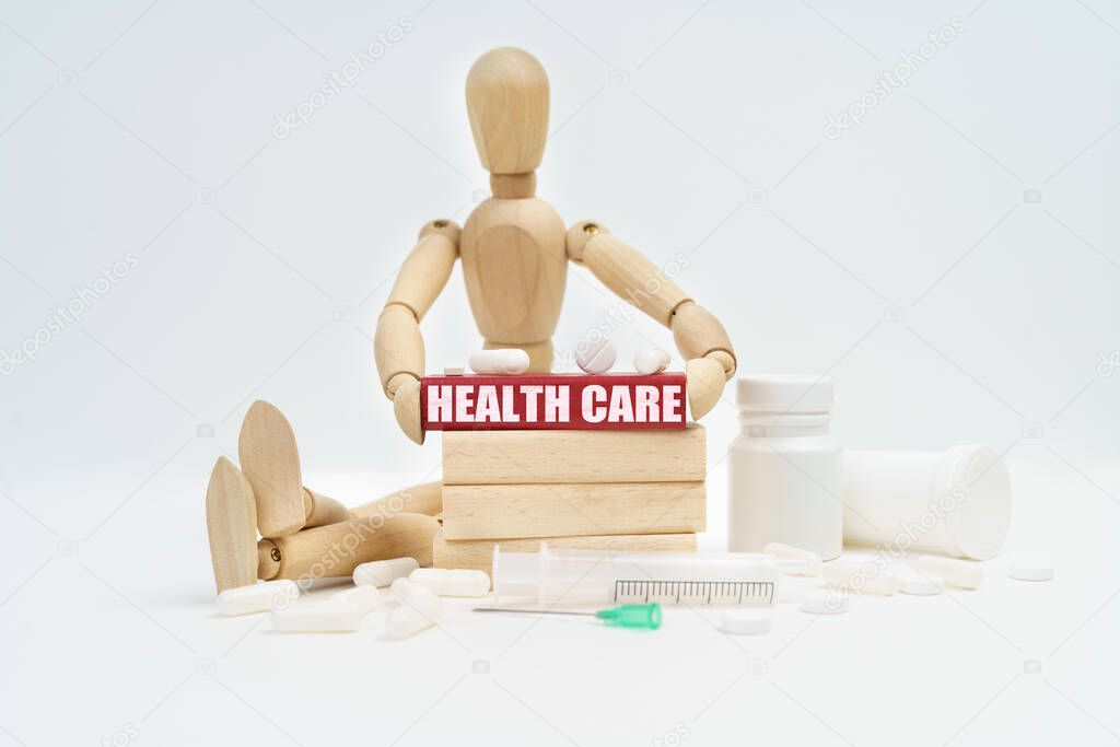 Medicine and healthcare concept. A figurine of a man sitting among pills lifts a red wooden block with an inscription HEALTH CARE from a wall of blocks.