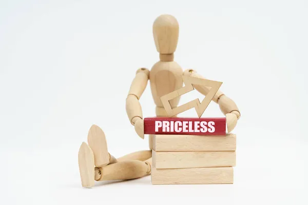 Business and Economics. On a white background, a wooden man sits near the wooden blocks, he holds in his hands a red block with the inscription - Priceless