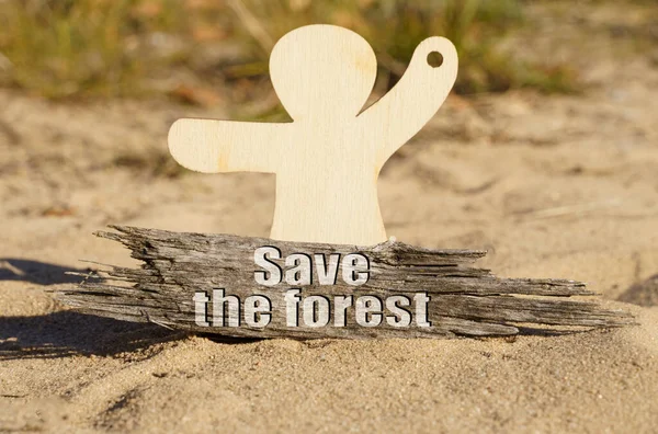 Op het zand bij het houten beeldje van een man staat een stuk hout met de inscriptie - Save the forest — Stockfoto