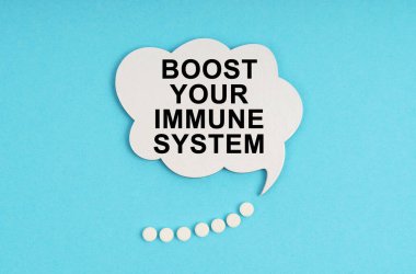 Mavi bir masada haplar ve üzerinde yazı olan beyaz bir tabak var. Bağışıklık sistemini güçlendir.