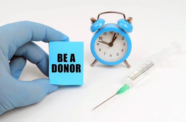 Sobre la mesa hay una jeringa y un despertador, en su mano un cuaderno con la inscripción - BE A DONOR —  Fotos de Stock