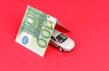 Araba sigortası konsepti. Garajda 100 avroluk bir banknotun içinde, bir araba. Kırmızı arkaplanda izole