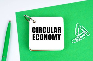 Beyaz bir yüzeyde yeşil bir kağıt, bir kalem ve üzerinde yuvarlak ekonomi yazan bir defter bulunur.