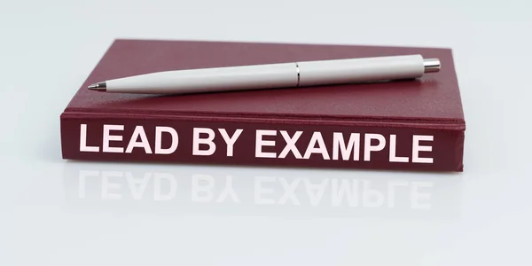 On a white surface lies a pen and a notebook with the inscription - LEAD BY EXAMPLE — 스톡 사진