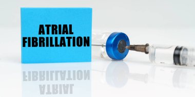 On a white reflective surface are a syringe, an injection and a blue plaque that says - Atrial Fibrillation