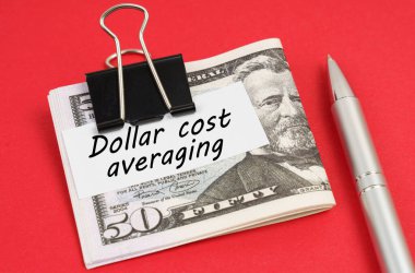 Finance and business concept. On a red background lies a pen and dollars clamped with a clip with the inscription on paper - Dollar cost averaging