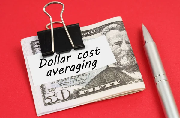 Finance and business concept. On a red background lies a pen and dollars clamped with a clip with the inscription on paper - Dollar cost averaging