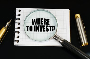 On a black surface there is a pen, a notebook and a magnifying glass. The inscription in the notebook - WHERE TO INVEST