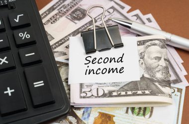 Finans ve iş konsepti. Kahverengi arka planda bir hesap makinesi ve bir şarjörde dolar ve kağıt üzerinde bir yazı bulunur - ikinci gelir