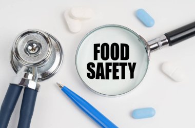 Tıp ve sağlık konsepti. Beyaz bir yüzeyde yalan hapları, bir kalem, bir steteskop ve üzerinde Gıda Güvenliği yazan bir büyüteç.