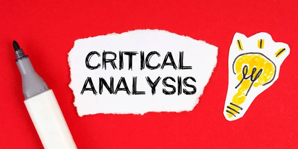 Kırmızı bir yüzeyde bir işaret, bir ampul çizimi ve üzerinde CRITICAL AnALYSIS yazan yırtık bir kağıt bulunur.