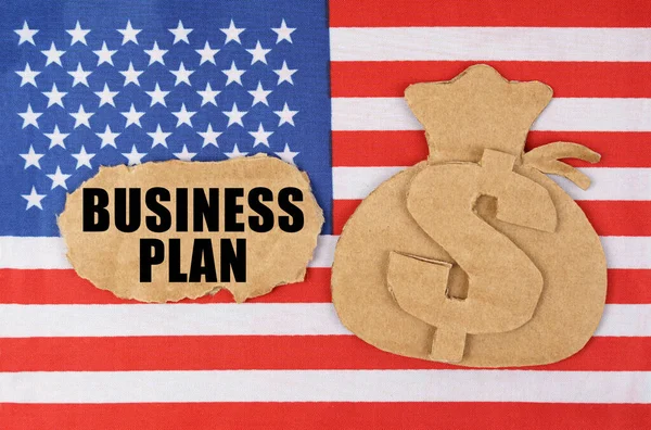På den amerikanska flaggan finns en pappfigur av en pengapåse och en kartong med inskriptionen - BUSINESS PLAN — Stockfoto
