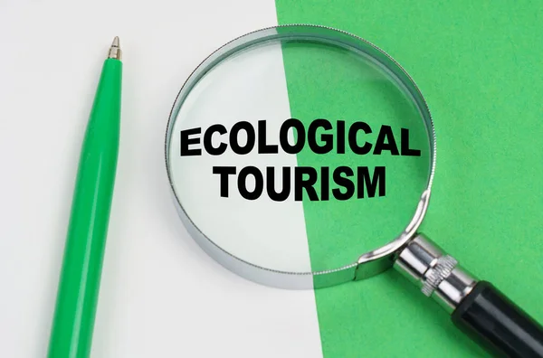 Auf weißgrünem Hintergrund liegt ein Stift und eine Lupe, in der sich die Aufschrift befindet - Ökotourismus — Stockfoto