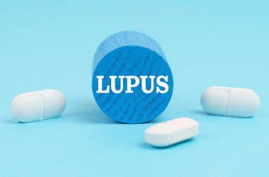 Tıp ve sağlık konsepti. Haplar mavi bir yüzeyde duruyor, üzerinde LUPUS yazan ahşap mavi bir silindir.
