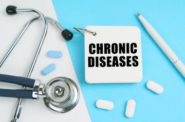 Tıbbi konsept. Beyaz ve mavi yüzeyde haplar, bir steteskop, bir kalem ve üzerinde CHRONIC DISEASES yazan bir not defteri var.