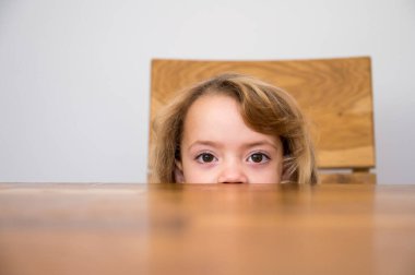 Cute blonde girl hiding half of her face under the wooden table.