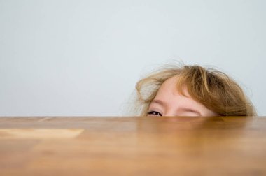 Cute blonde girl hiding half of her face under the wooden table.
