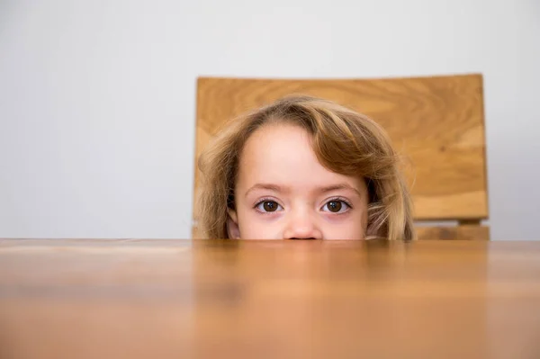 Cute blonde girl hiding half of her face under the wooden table. — Stockfoto