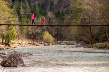 Anne ve kızı nehrin karşısındaki asma köprüden geçiyorlar. Soca Vadisi, Slovenya.