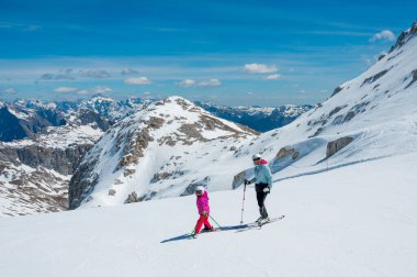 Anne ve kızı dağ tepesindeki bir kayak merkezinde ilk adımlarını atarak eğleniyor ve kayak öğreniyor. Kış boyunca aktif çocukluk.