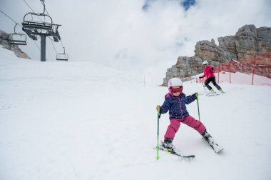 Anne ve kızı dağ tepesindeki bir kayak merkezinde ilk adımlarını atarak eğleniyor ve kayak öğreniyor. Kış boyunca aktif çocukluk.