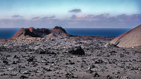 A moon-like, unreal landscape in the heart of the volcanic island of Lanzarote, in the Fire Mountains or Timanfaya Mountains. Black lava stones, a crater opening of a small volcanic cone in the evening light. Behind the dark blue Atlantic and a few g