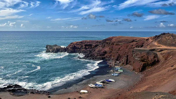 Lanzarote volkanik adasındaki El Golfo balıkçı köyünün yakınındaki karanlık kumlu sahilde küçük balıkçı tekneleri olan küçük bir koy. Çevredeki kayalar kırmızı ve siyah, bitki örtüsü falan yok. Koyu mavi Atlantik ve üzerinde mavi gökyüzü 