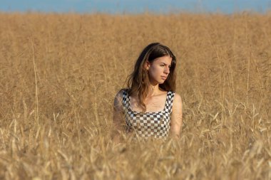 Güneşli bir günde, buğday tarlasında yarı çıplak duran ve başka tarafa bakan genç bir kadın.