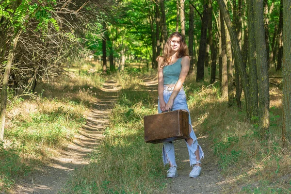 森の未舗装の道に立つ若い女性と古い茶色のスーツケースを保持 — ストック写真