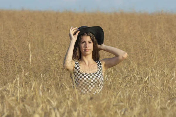 黒い帽子をかぶった若い女性が立ち カメラの半分を見て 晴れた日に小麦畑に覆われて — ストック写真
