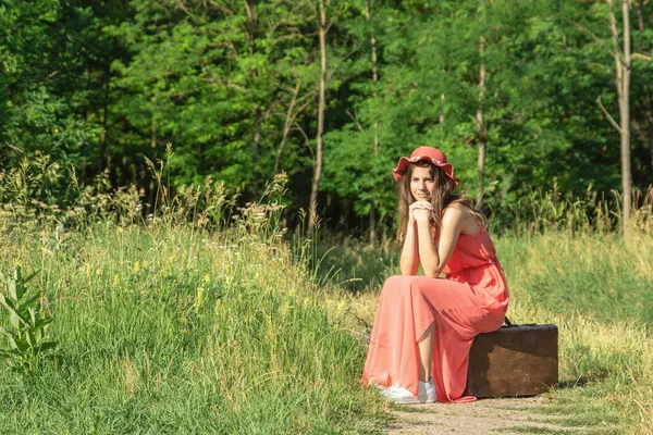 若いです女性で赤いドレスとともに帽子座っています古い茶色のスーツケースで森林未舗装の道路 — ストック写真