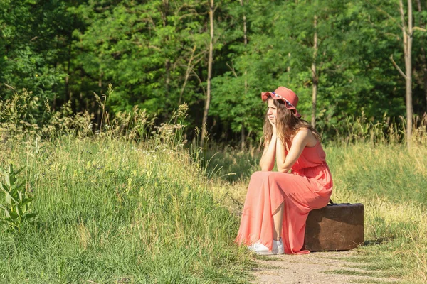 若いです女性で赤いドレスとともに帽子座っています古い茶色のスーツケースで森林未舗装の道路 — ストック写真