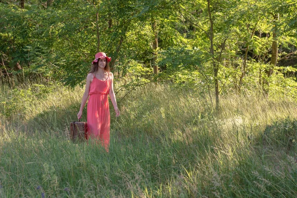 若いです女性で赤いドレスで歩くで森林未舗装の道路とともに古い茶色のスーツケース — ストック写真