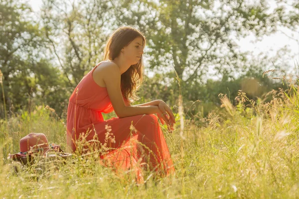 若い女性で赤いドレススーツケースに座っているとバックライト付きの森の中でカメラを見て — ストック写真