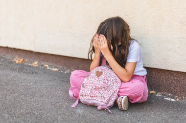 Küçük esmer kız elleriyle yüzünü gizliyor, yalnız oturuyor ve üzgün hissediyor.