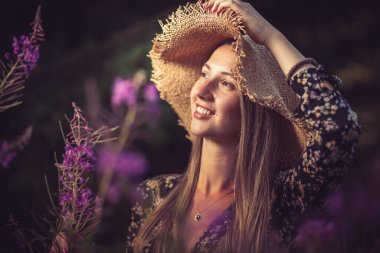Etrafı mor çiçeklerle çevrili yaz şapkalı genç bir kadın.