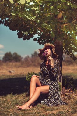 Yazlık şapkalı genç bir kadın, ağacın yanında oturuyor.